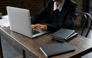 Close-up of a person in a suit typing on a laptop at a wooden desk with notebooks.