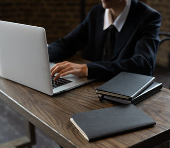 Close-up of a person in a suit typing on a laptop at a wooden desk with notebooks.