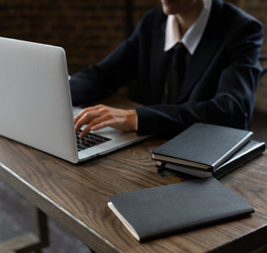 Close-up of a person in a suit typing on a laptop at a wooden desk with notebooks.