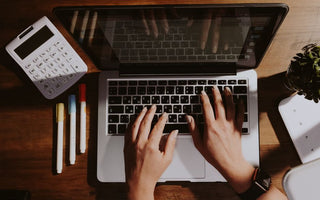 Hands typing on a laptop with a calculator and pens nearby on a wooden table.