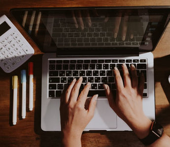 Hands typing on a laptop with a calculator and pens nearby on a wooden table.