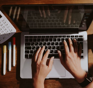 Hands typing on a laptop with a calculator and pens nearby on a wooden table.