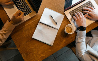 Overhead shot of coworkers using laptops with notes and coffee at a wooden desk.