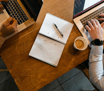 Overhead shot of coworkers using laptops with notes and coffee at a wooden desk.