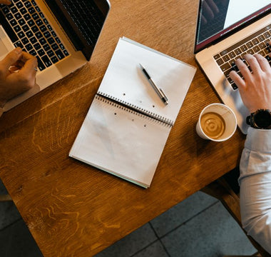Overhead shot of coworkers using laptops with notes and coffee at a wooden desk.