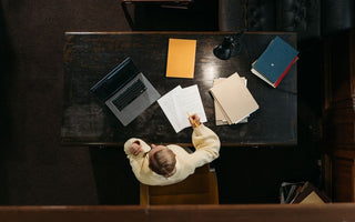 Person in yellow sweater working at a wooden desk with documents, folders, and a laptop.
