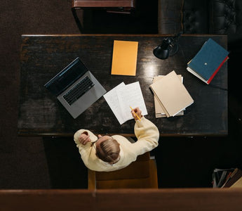 Person in yellow sweater working at a wooden desk with documents, folders, and a laptop.