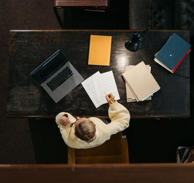 Person in yellow sweater working at a wooden desk with documents, folders, and a laptop.